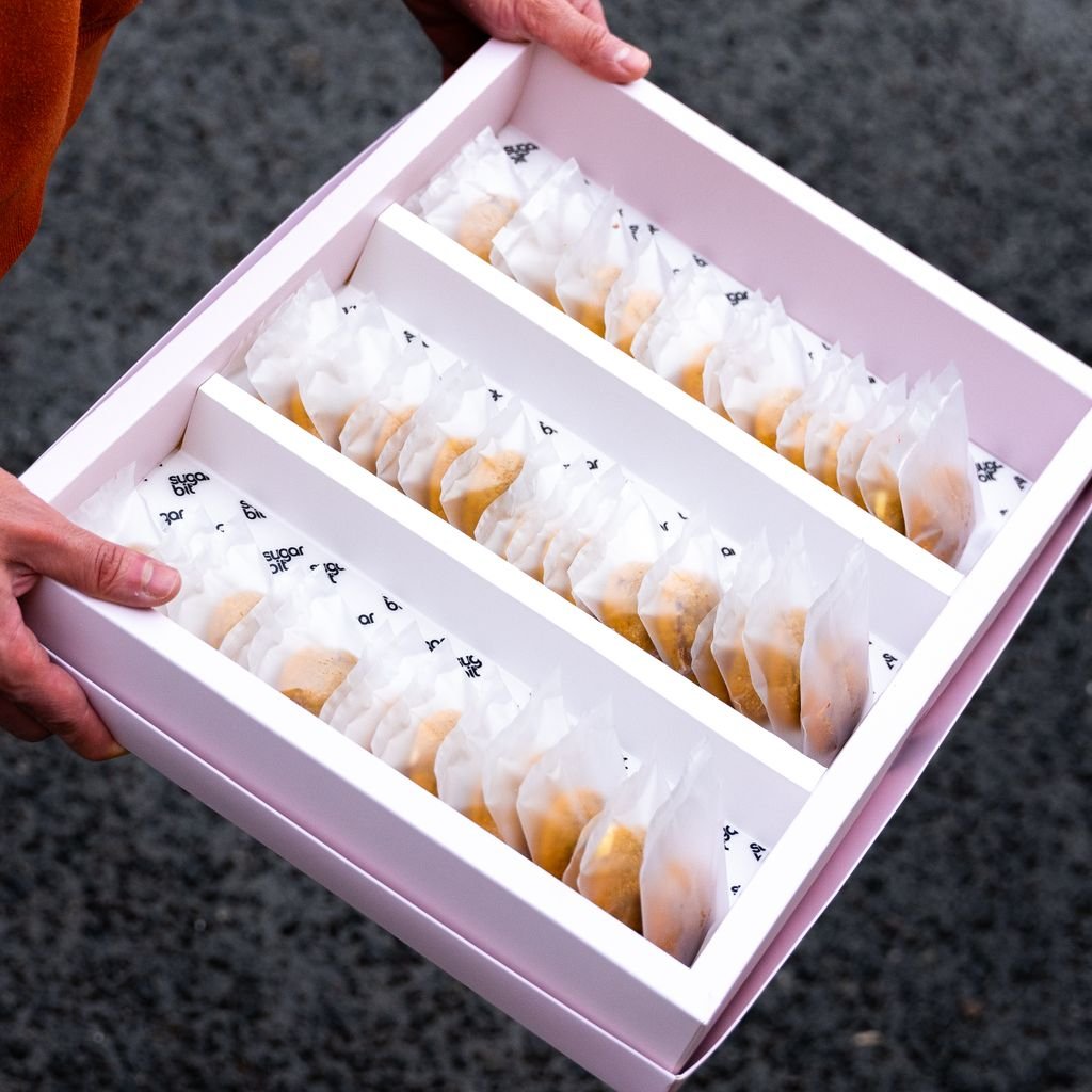 A man holds a pink box filled with individually wrapped cookies on a gray background.
