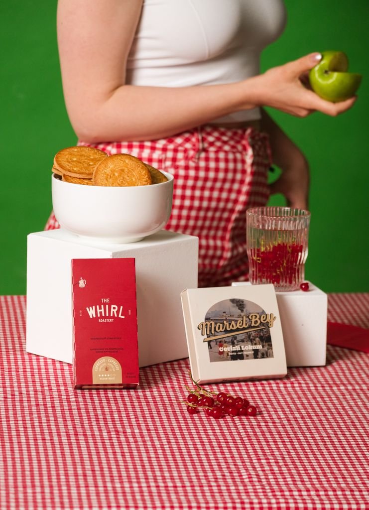 A person in red gingham holds a green apple beside biscuits and packaged food on a table.