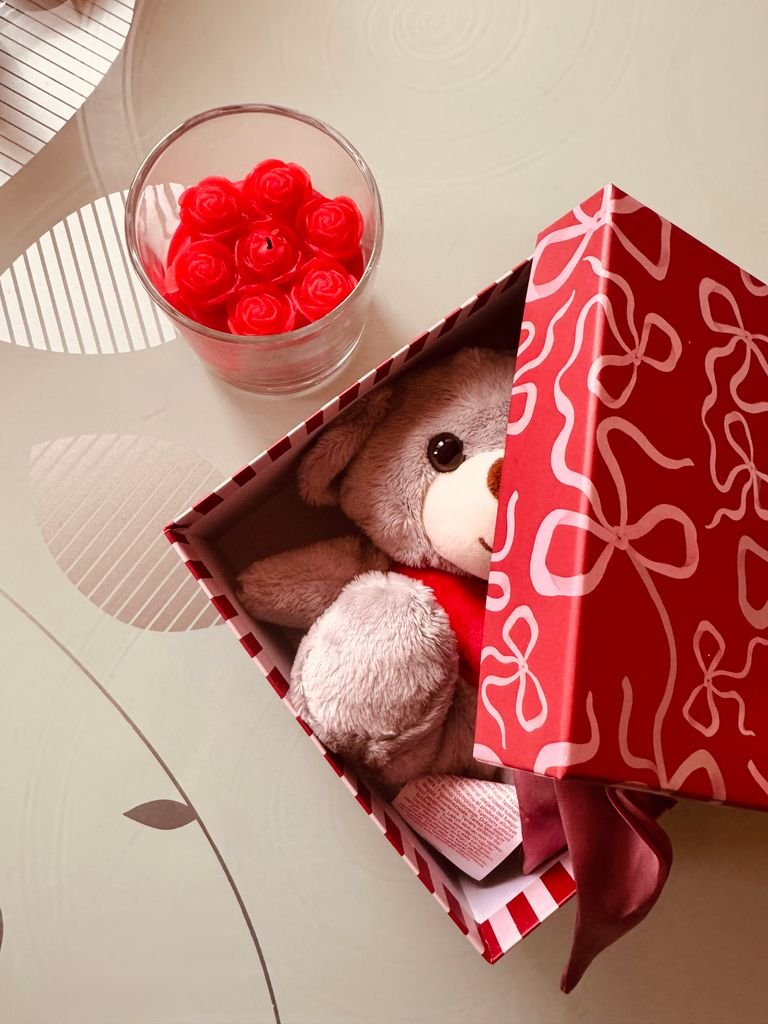 A teddy bear rests in a decorated gift box beside a cup of decorative red roses.