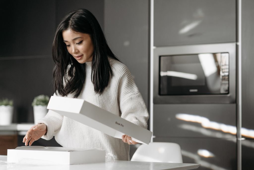 An asian woman excitedly unpacks a delivery box in her modern kitchen.